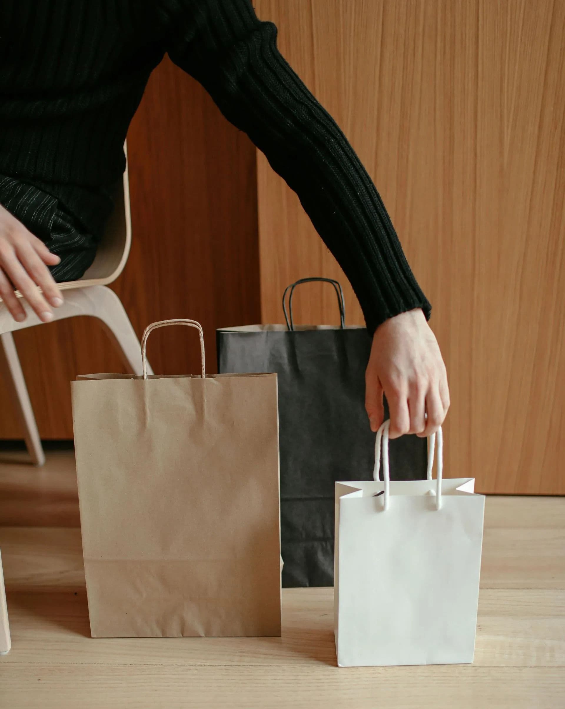 A person in a black sweater reaches for a white paper bag on a wooden floor. Nearby, a brown and a black paper bag are on the floor. Calm and minimalistic setting.