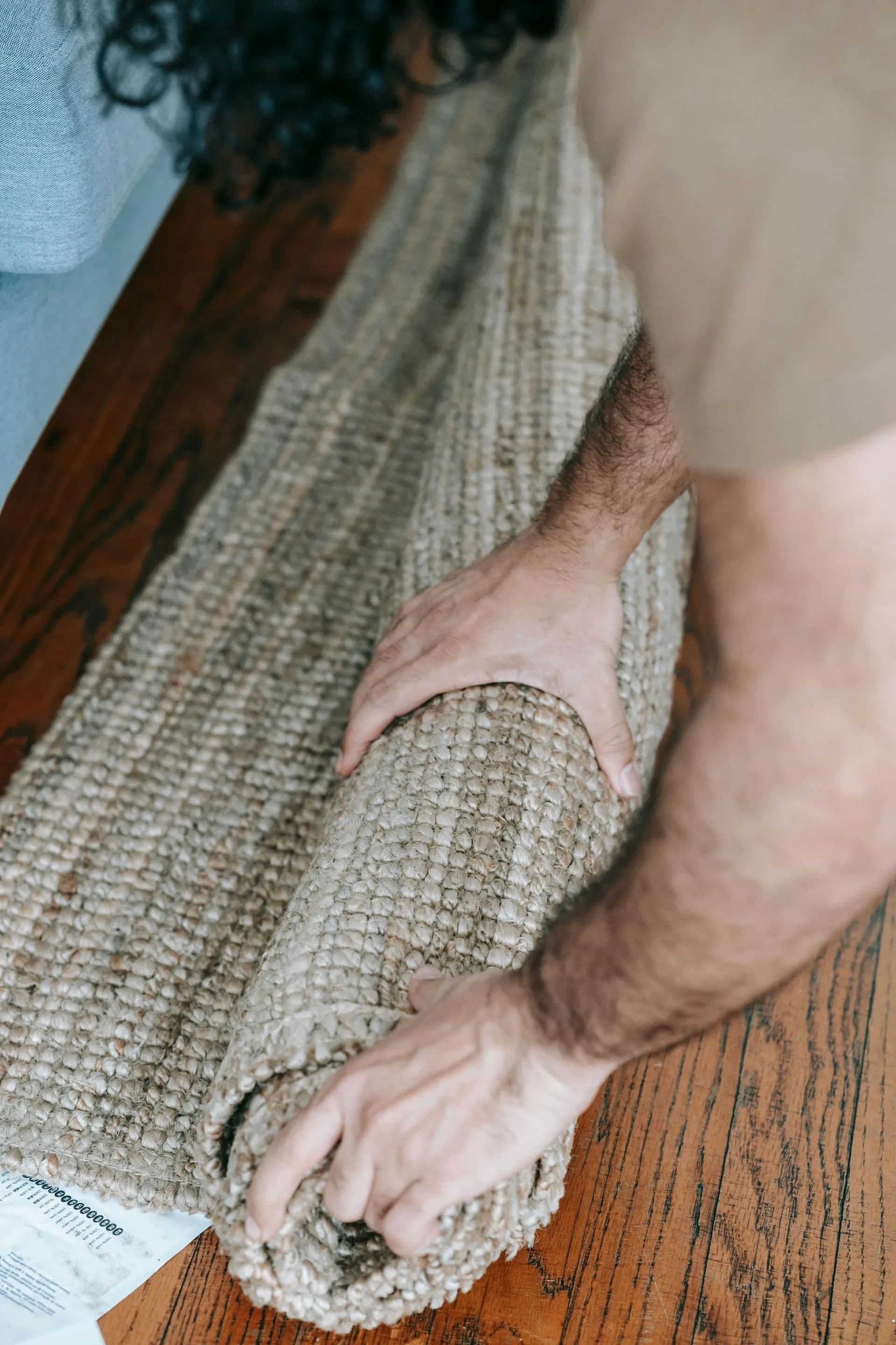 Person rolling a textured, beige rug on a wooden floor, with focused hands and forearms. A sheet of paper is partially visible underneath. Calm, domestic setting.