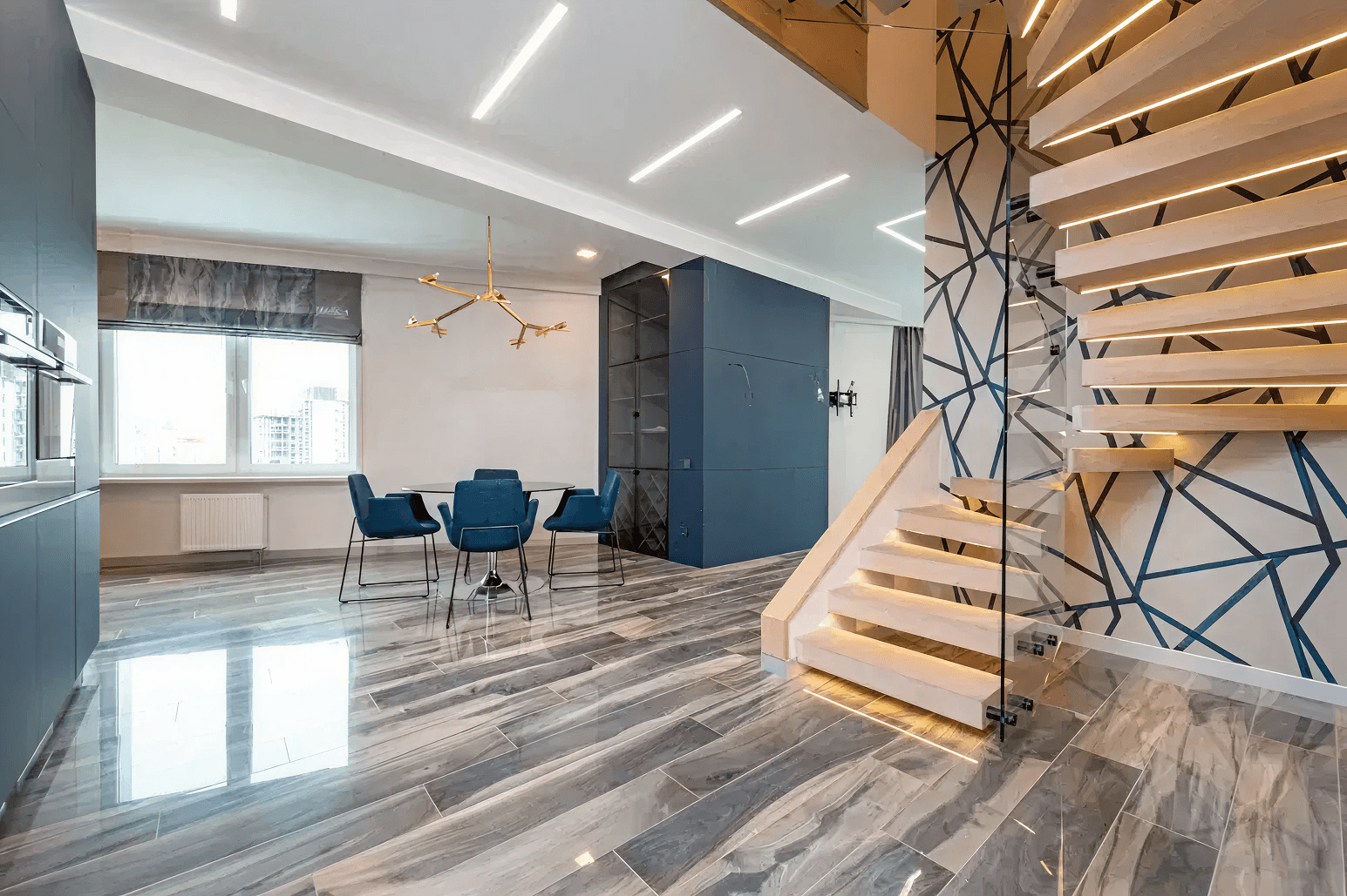 Modern interior with geometric staircase, blue-gray walls, and a sleek gray floor. A gold chandelier hangs above a table with blue chairs, creating a stylish, elegant atmosphere.