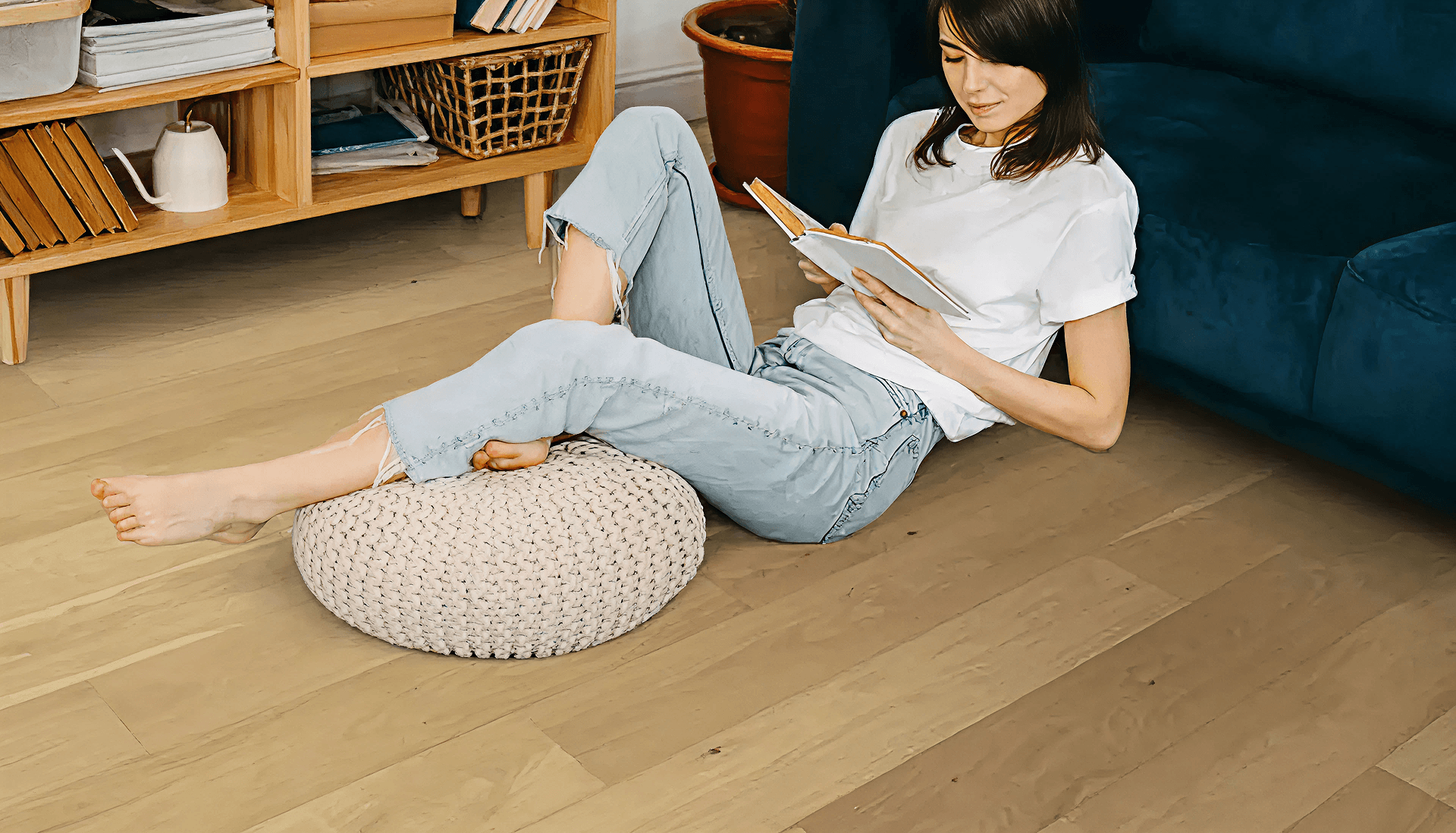 Woman in casual jeans and a white t-shirt reads a book while reclining on a knitted pouf on a wooden floor, near a book-filled shelf, conveying relaxation.