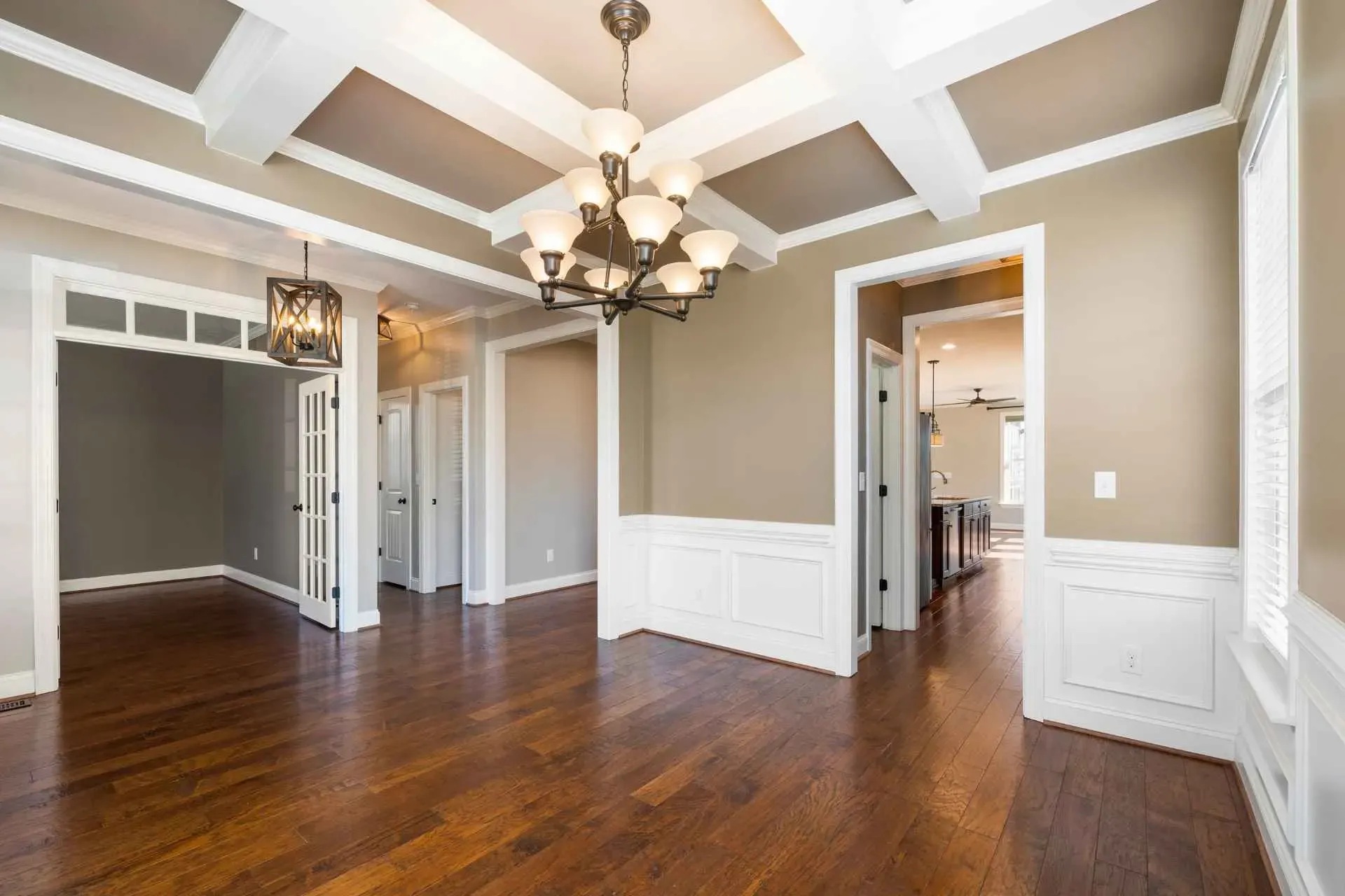 Elegant, empty dining room with warm wood floors, coffered ceiling, dual chandeliers, and beige walls with white wainscoting, leading to a kitchen.