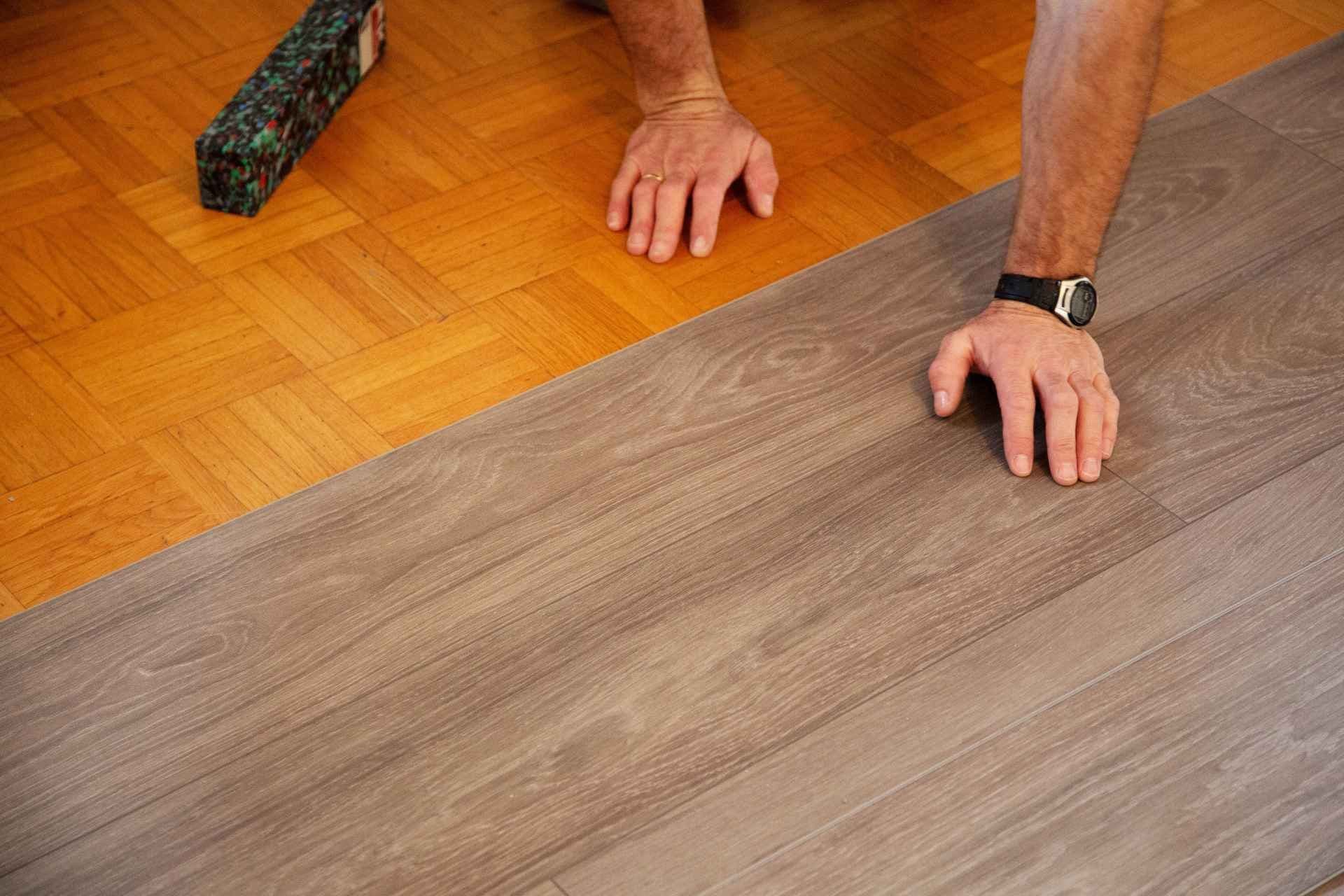 Two hands are installing gray laminate flooring over an old brown parquet floor. A box of materials rests nearby, indicating a home renovation project.