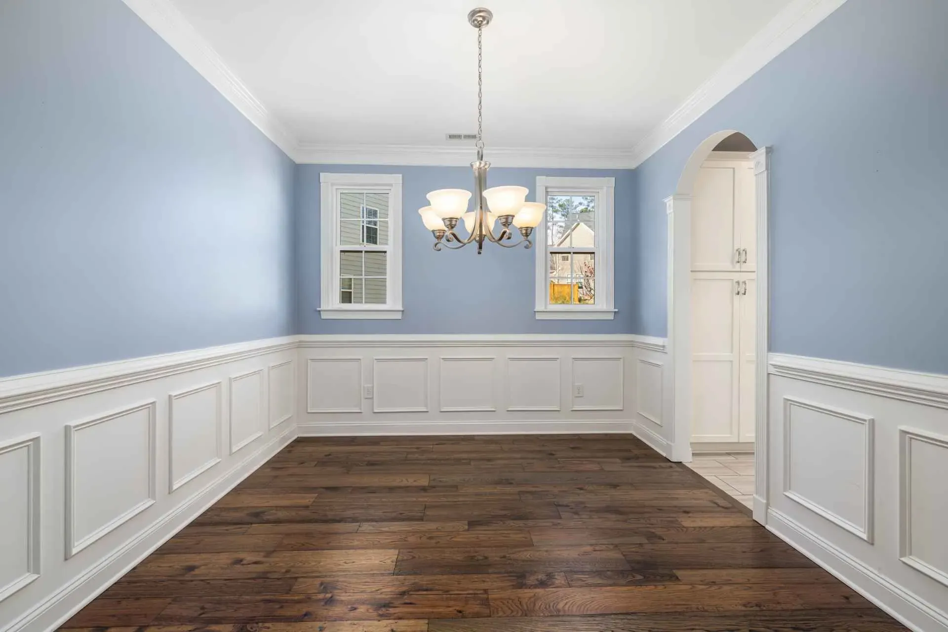 A bright dining room with light blue walls, white wainscoting, dark wood flooring, a five-light chandelier, and two windows. Tone is elegant and serene.