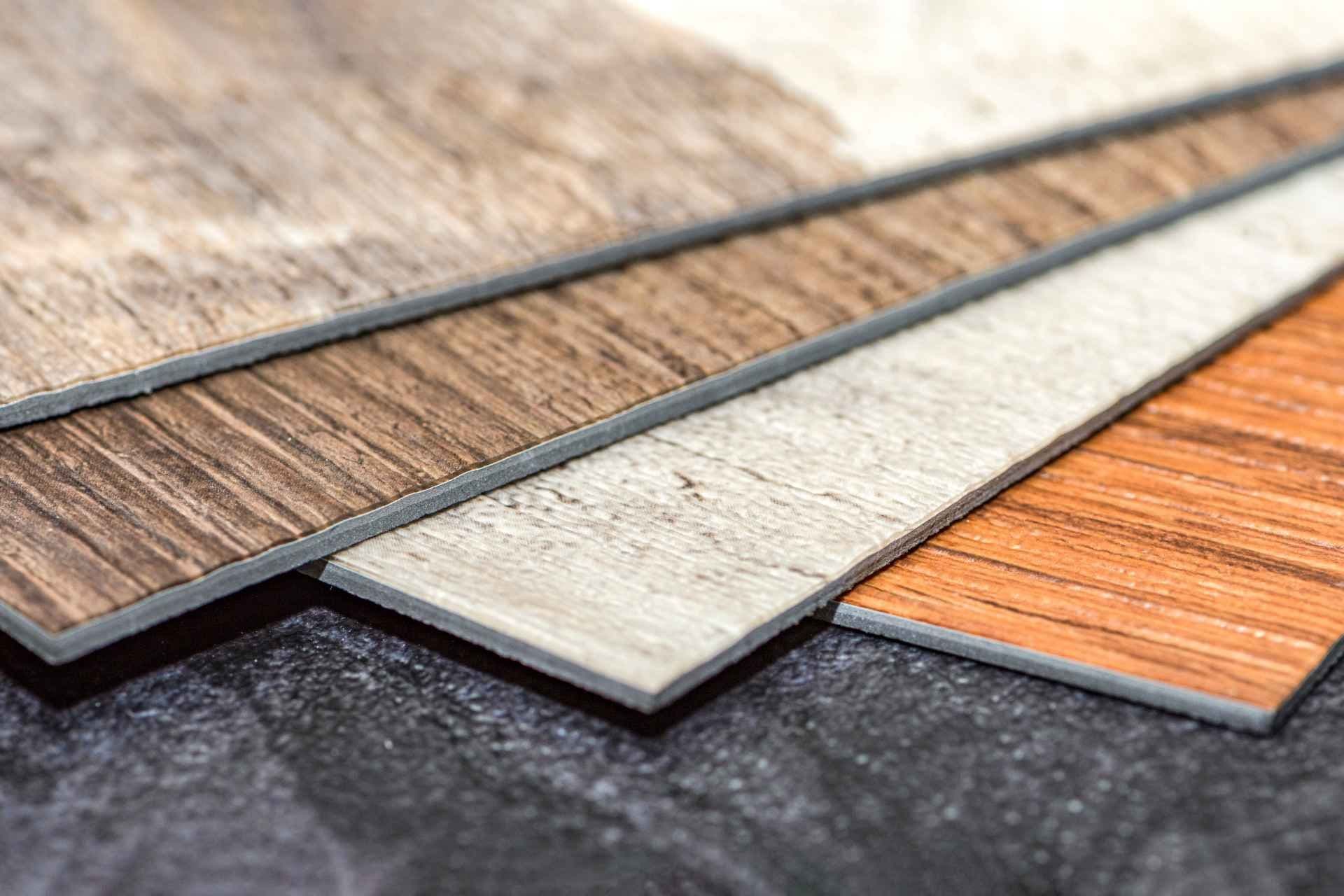 Close-up of four overlapping vinyl flooring samples in shades of brown, gray, and orange. They are layered on a dark textured surface.