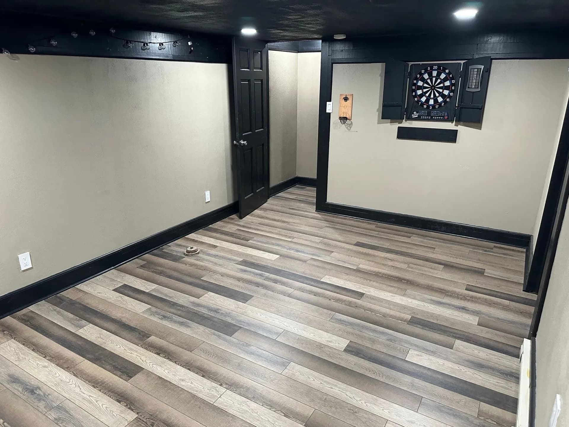 Small room with beige walls and dark trim, featuring a light wood floor. A dartboard is mounted on the right wall. The room has a cozy, game-room feel.