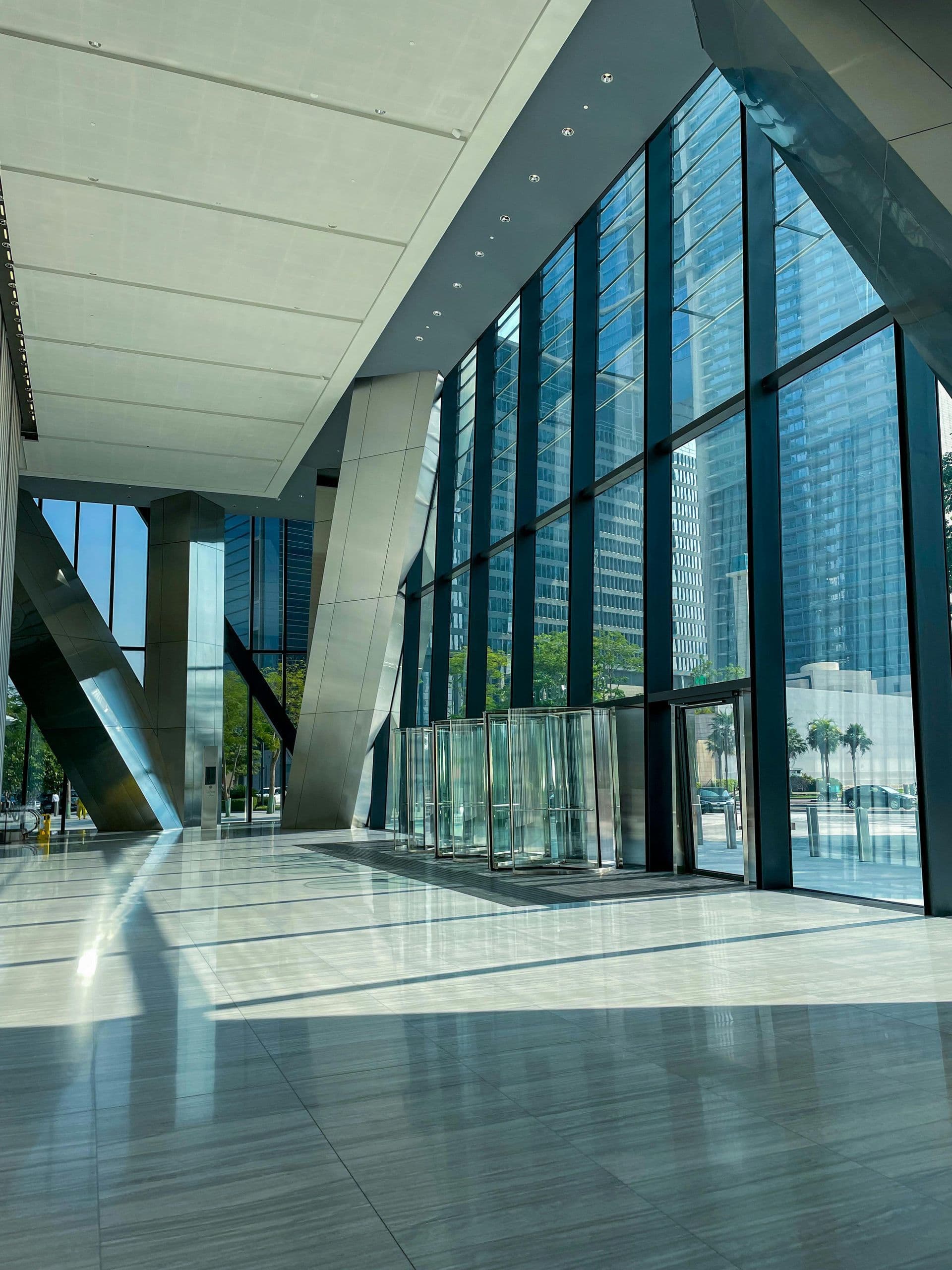 Modern building lobby with tall glass windows allowing abundant sunlight. Sleek metal beams and polished floors create a spacious, airy atmosphere.