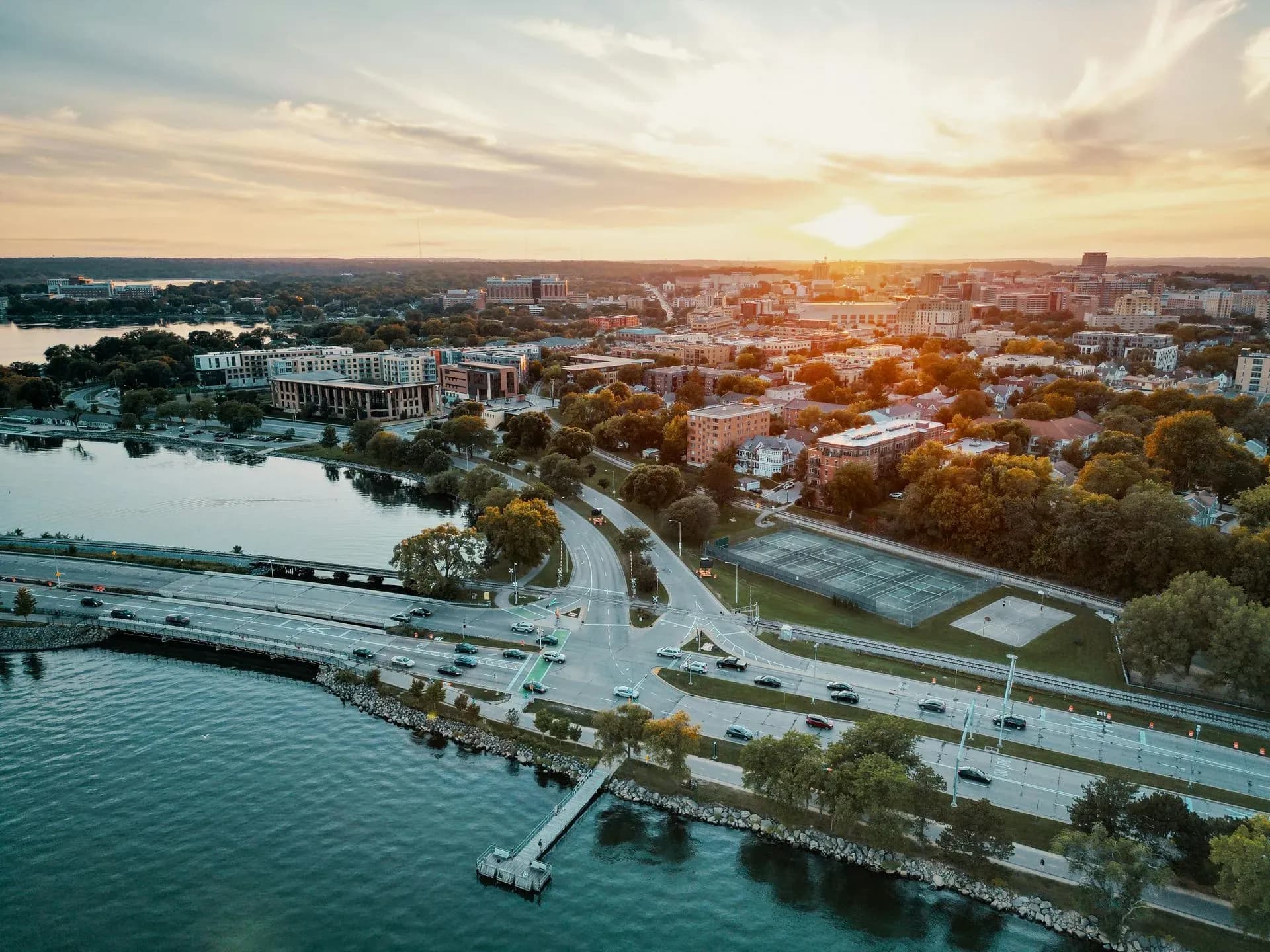 Aerial view of a cityscape at sunset, overlooking a waterfront with roads, trees, and buildings. The sky is vibrant with soft clouds and a warm glow.
