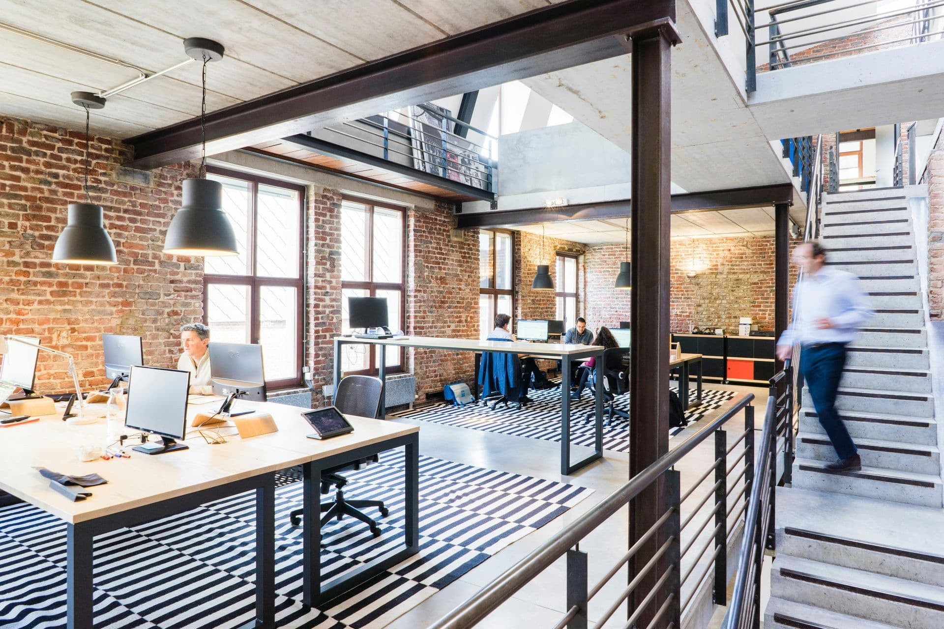 Open-plan office with brick walls and natural light. Three people work at desks with computers, while another walks down the stairs. Modern, dynamic atmosphere.