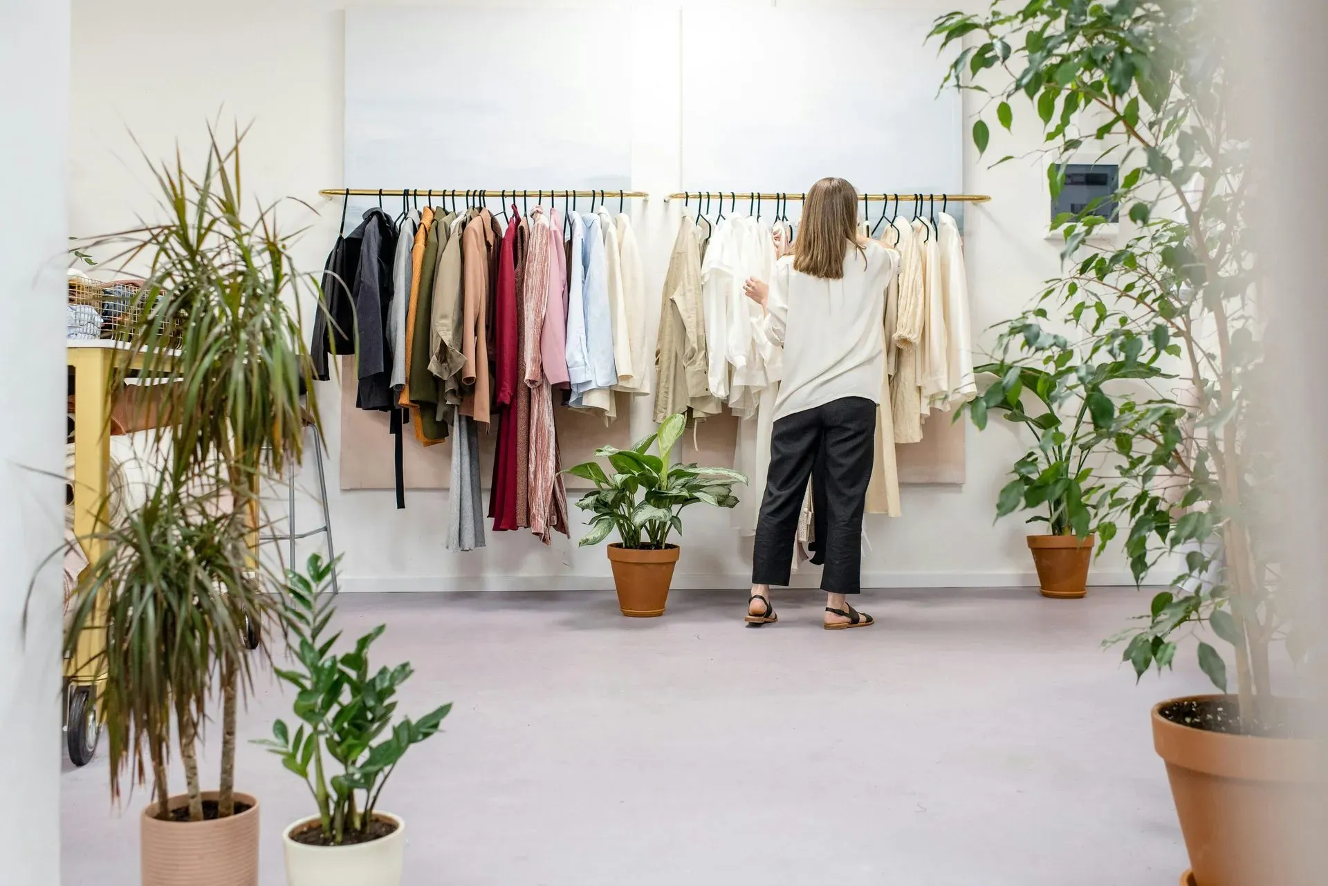A woman in a clothing store browses through racks of colorful garments. The space is bright and airy, with potted plants adding a natural touch.