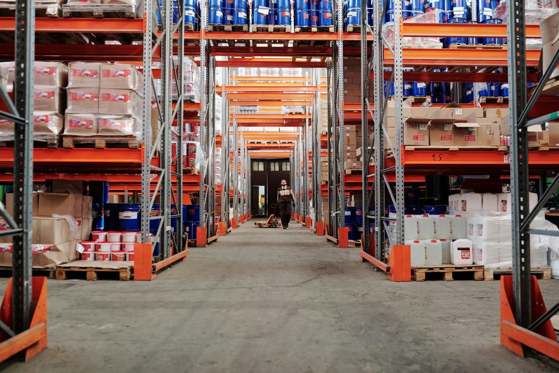 Wide warehouse aisle with orange shelves stacked with boxes and barrels. A person wearing a mask and apron walks down the aisle, conveying a sense of industry and focus.