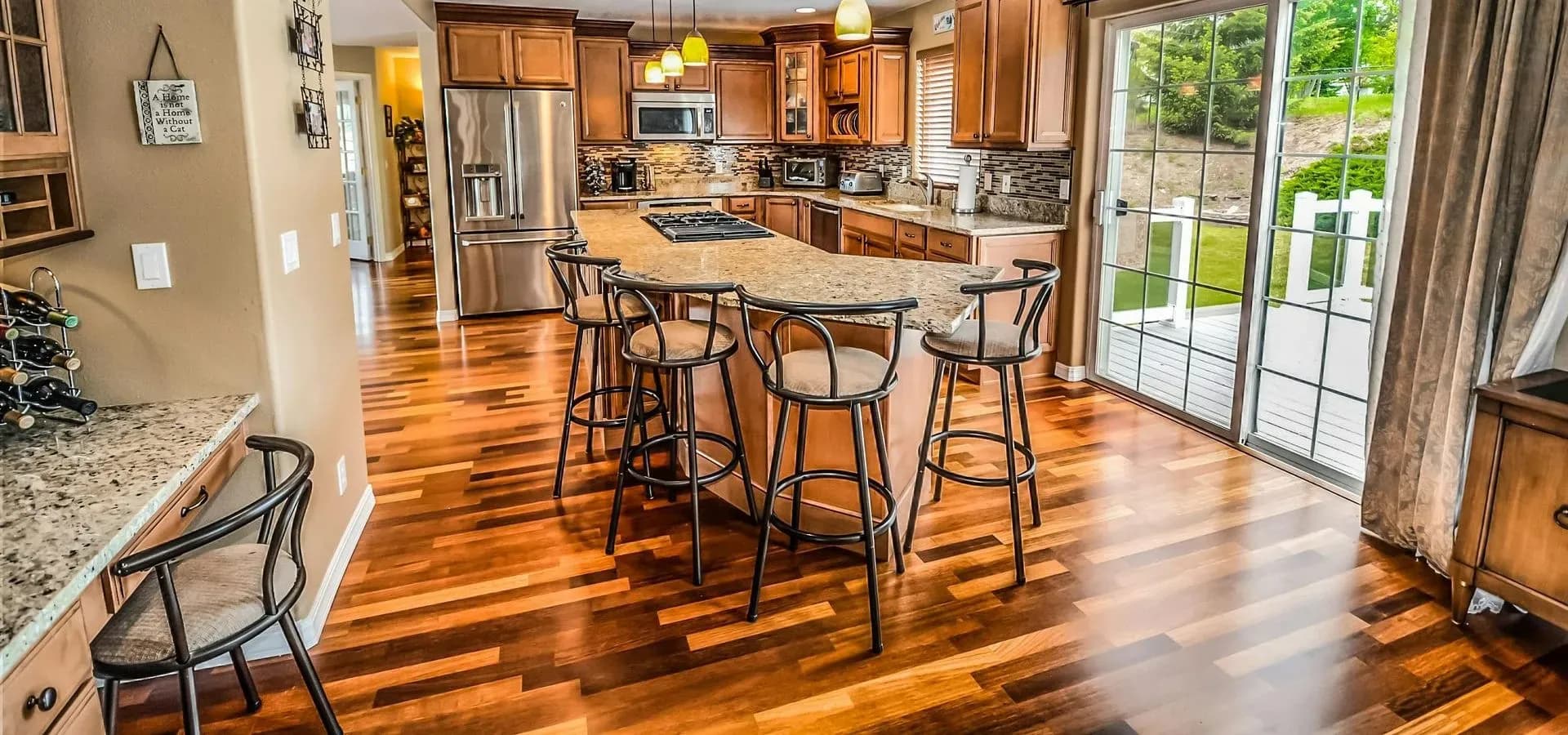 Spacious kitchen with wooden flooring, an island with bar stools, granite countertops, pendant lights, and a view of a patio through sliding glass doors.