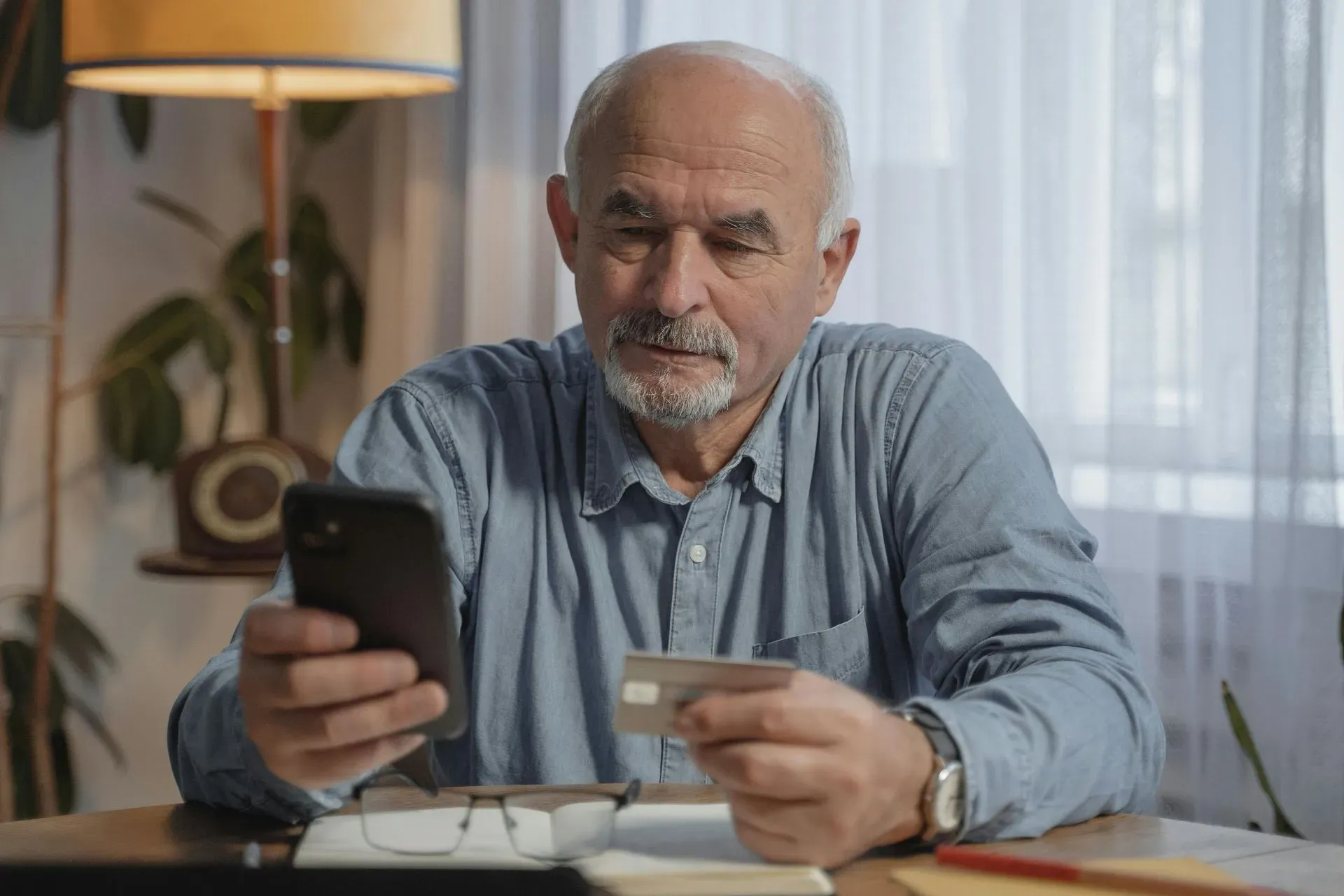 An elderly man in a denim shirt looks thoughtfully at a smartphone while holding a credit card. He's seated at a table with a lamp and houseplant.