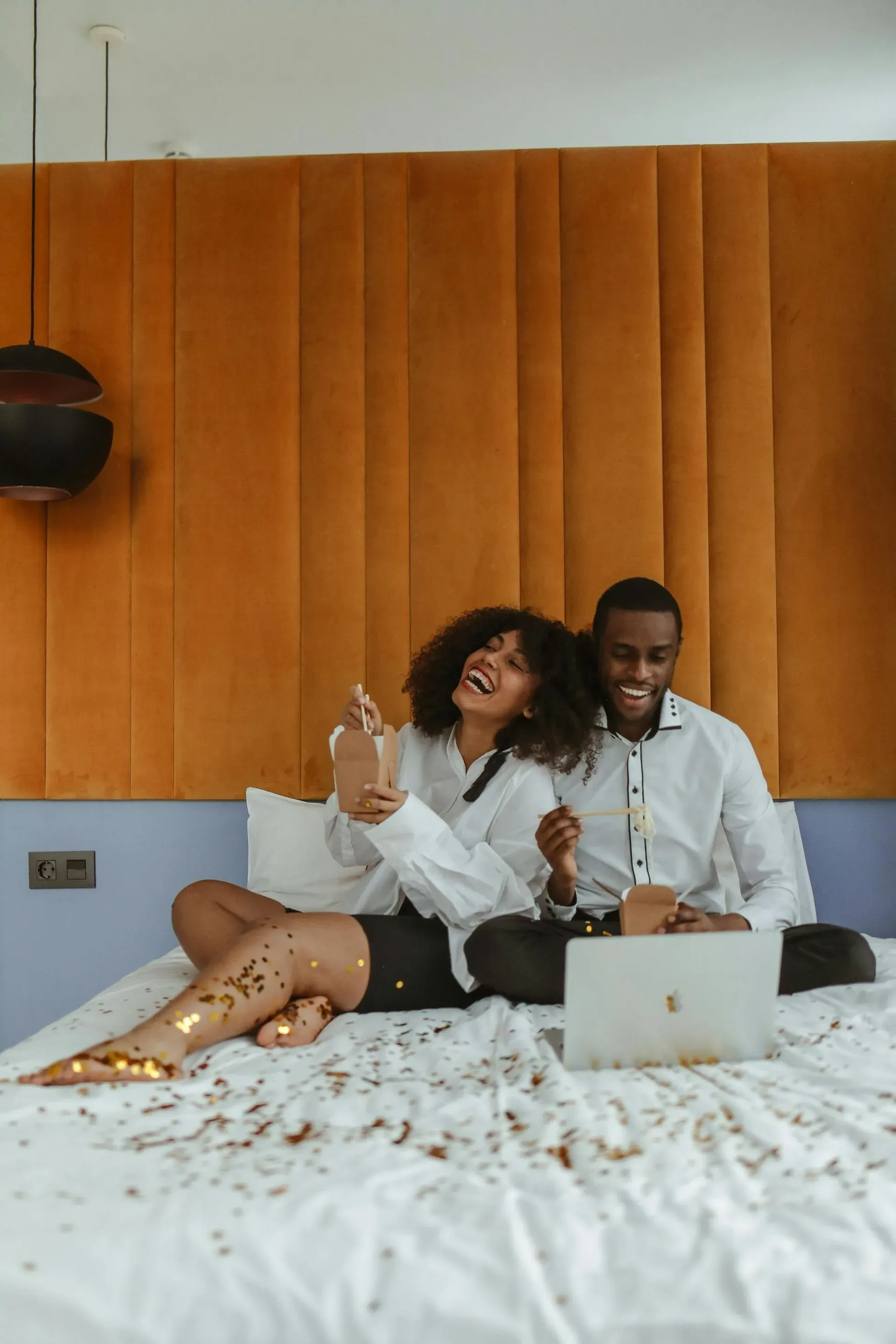 A joyful couple in white shirts sits on a bed, laughing and watching a laptop. Gold confetti is scattered around, creating a festive atmosphere.