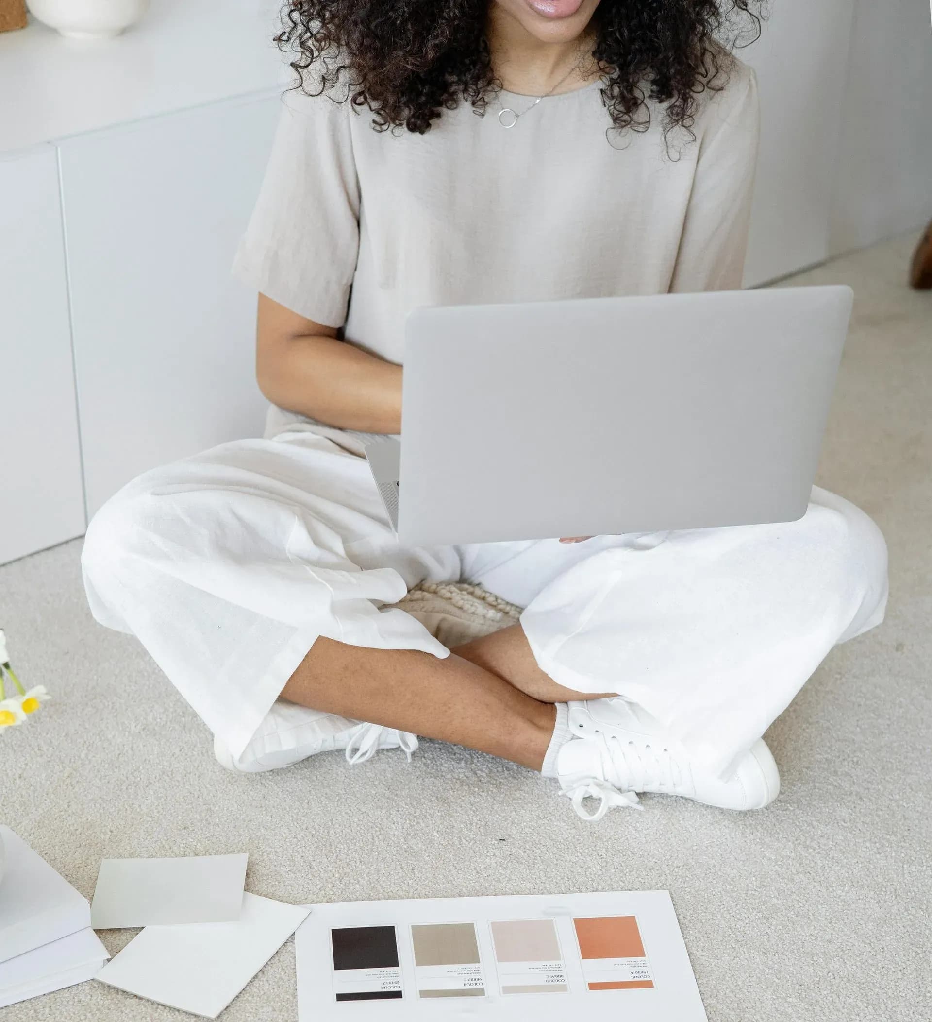 A woman with curly hair sits cross-legged on a carpeted floor, using a laptop. She wears a beige top and white pants. Paint swatches lie nearby.
