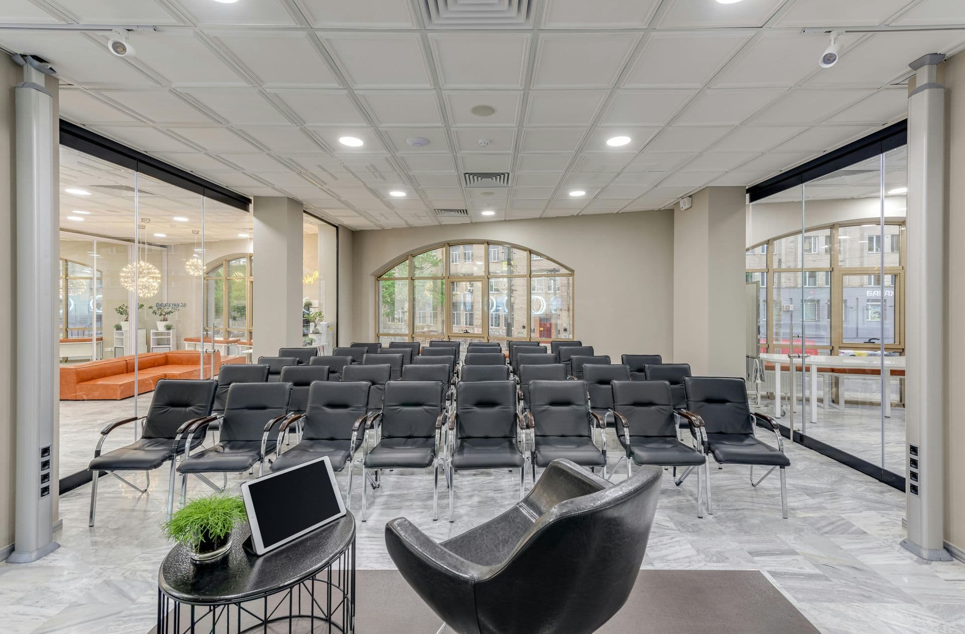 Modern conference room with rows of black chairs facing a speaker's area. Glass walls reveal an adjacent room with orange seating. Bright, professional ambiance.