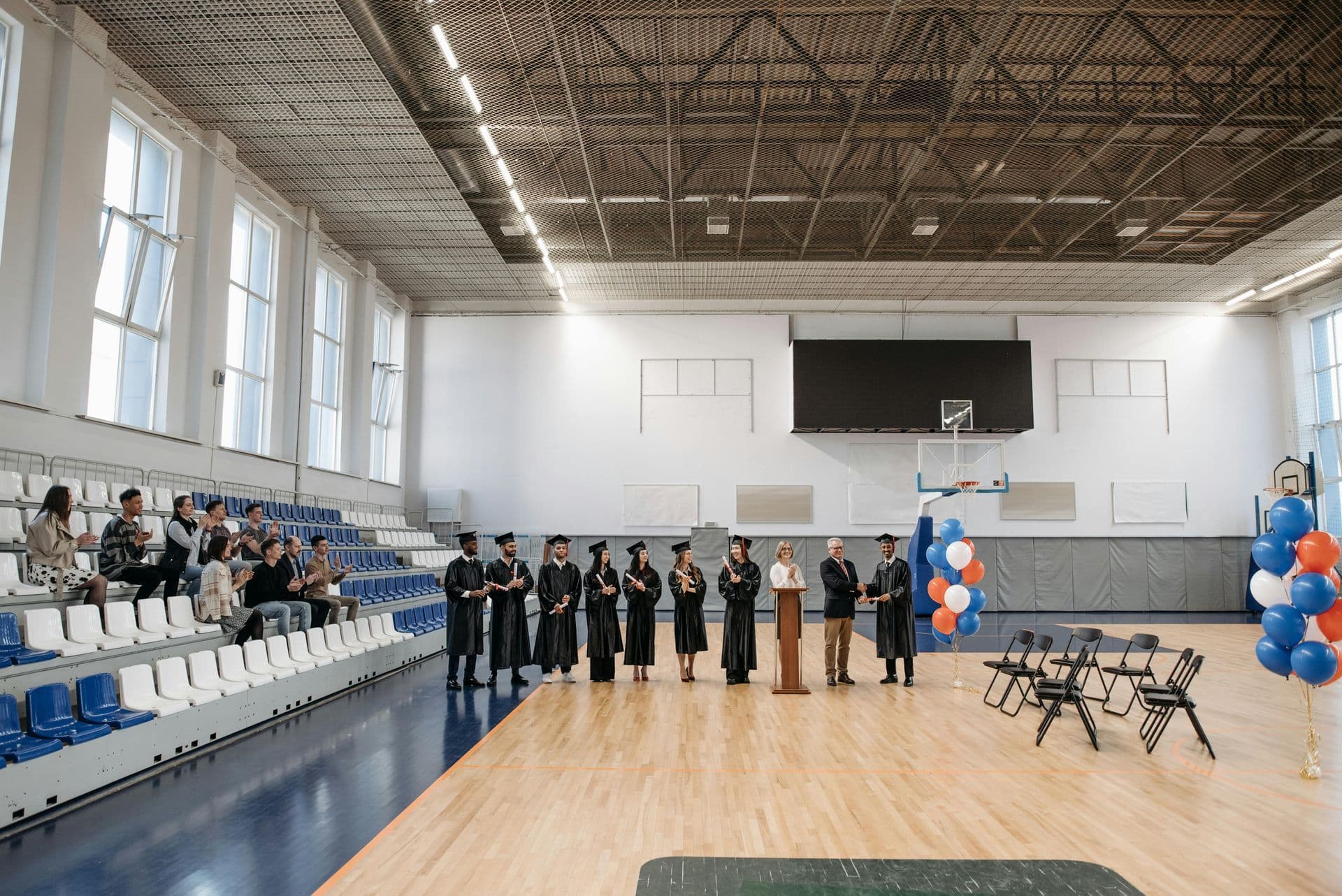 Graduation ceremony in a gym, featuring graduates in caps and gowns lined up beside a podium. Spectators sit in bleachers, surrounded by balloons.