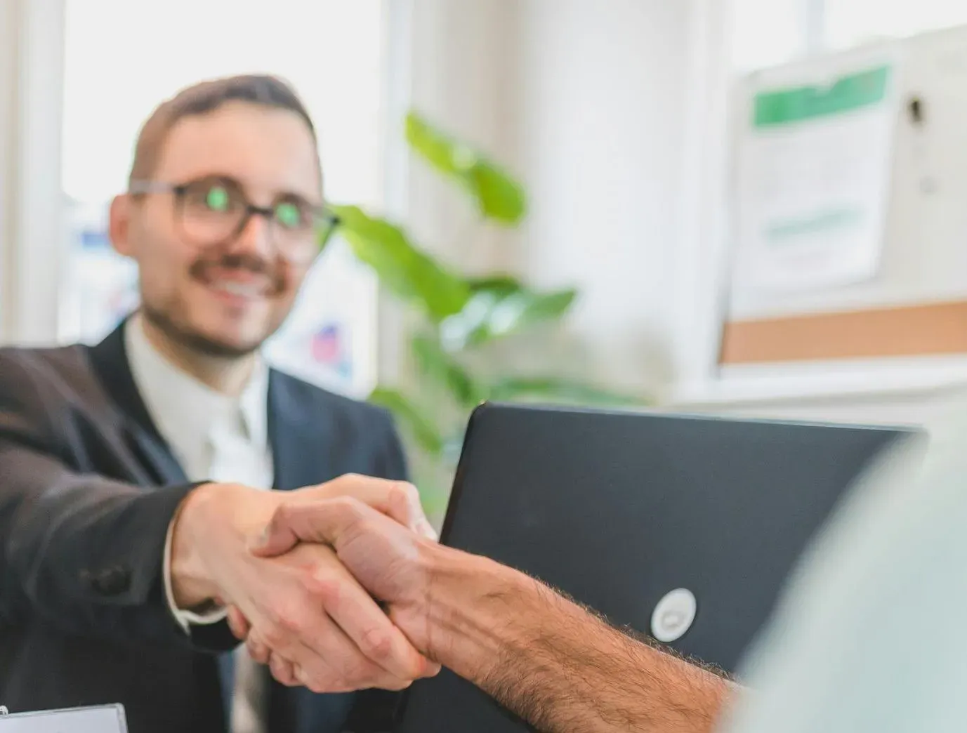 A man in a suit and glasses smiles warmly while shaking hands with another individual over a laptop. A plant and bright office setting suggest professionalism.