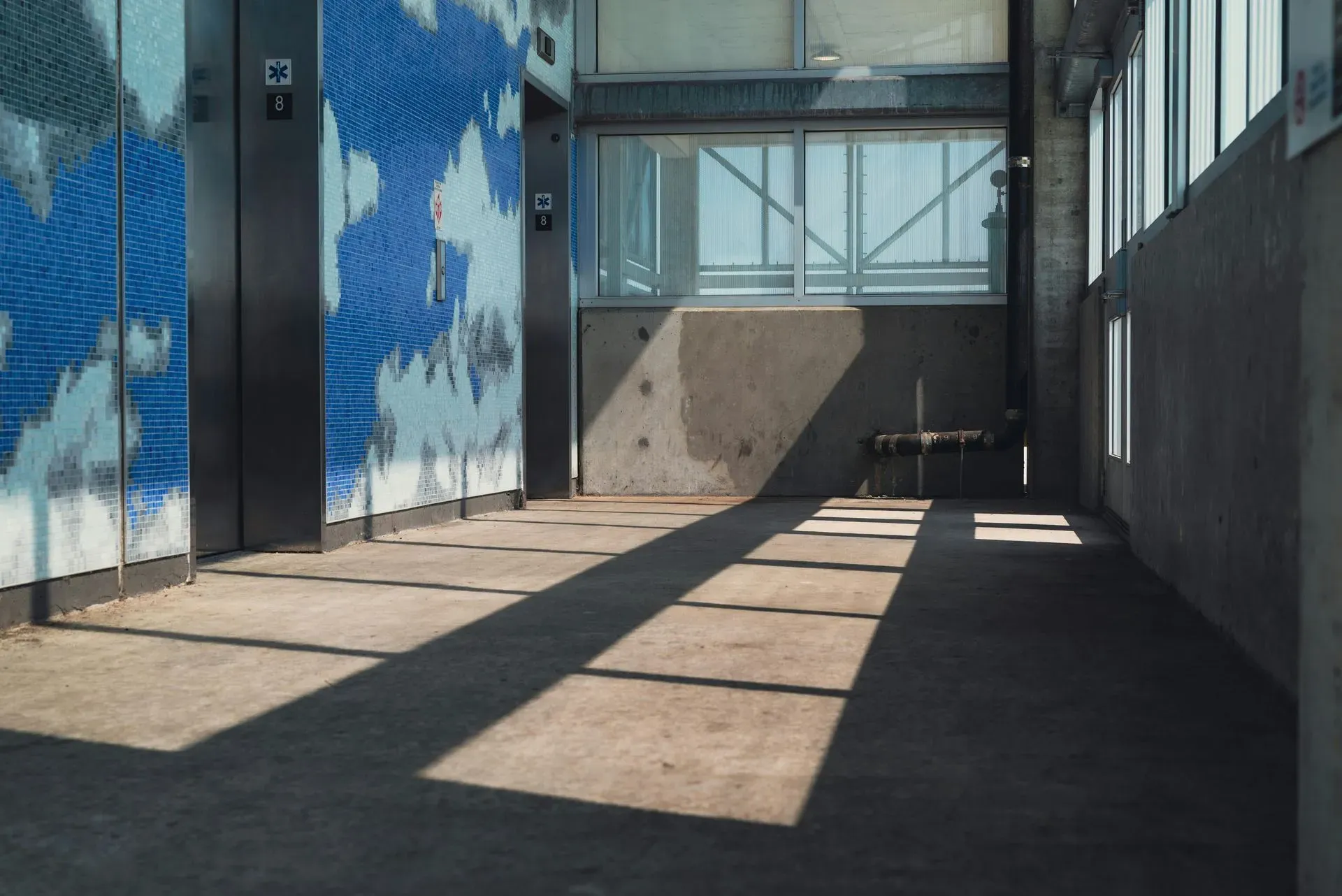 Sunlit corridor with shadowed grid patterns on concrete. Blue mosaic wall with cloud design on the left, large windows on the right. Quiet, serene ambiance.