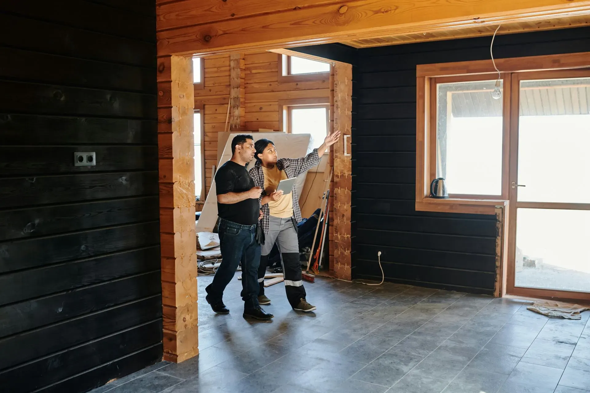 Two men stand in a partially renovated room with wooden walls and large windows. One gestures toward the ceiling, discussing the construction progress.