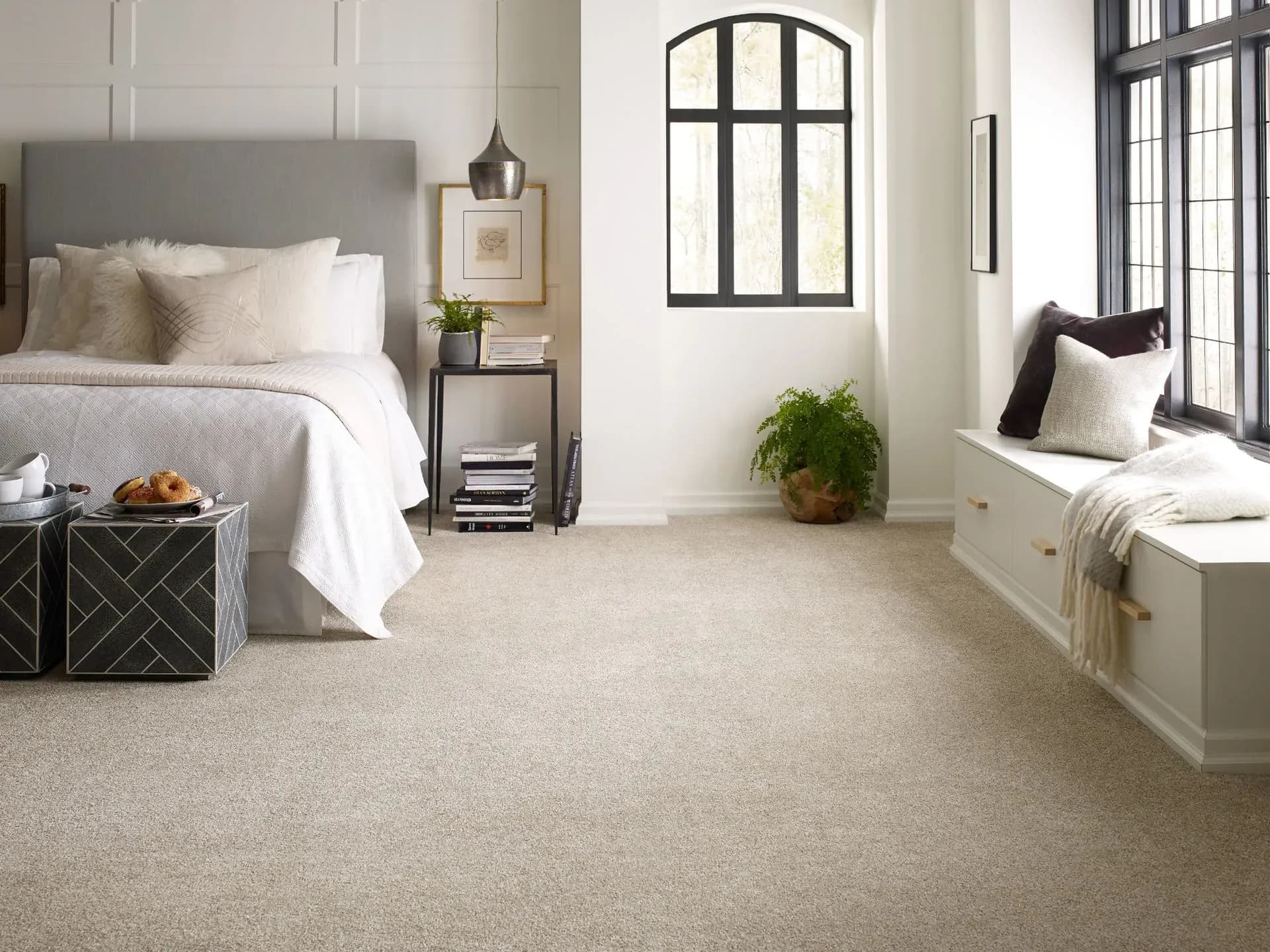 Cozy bedroom with a grey upholstered bed, white bedding, and a window seat with cushions. Two modern side tables hold breakfast pastries. Bright and inviting.