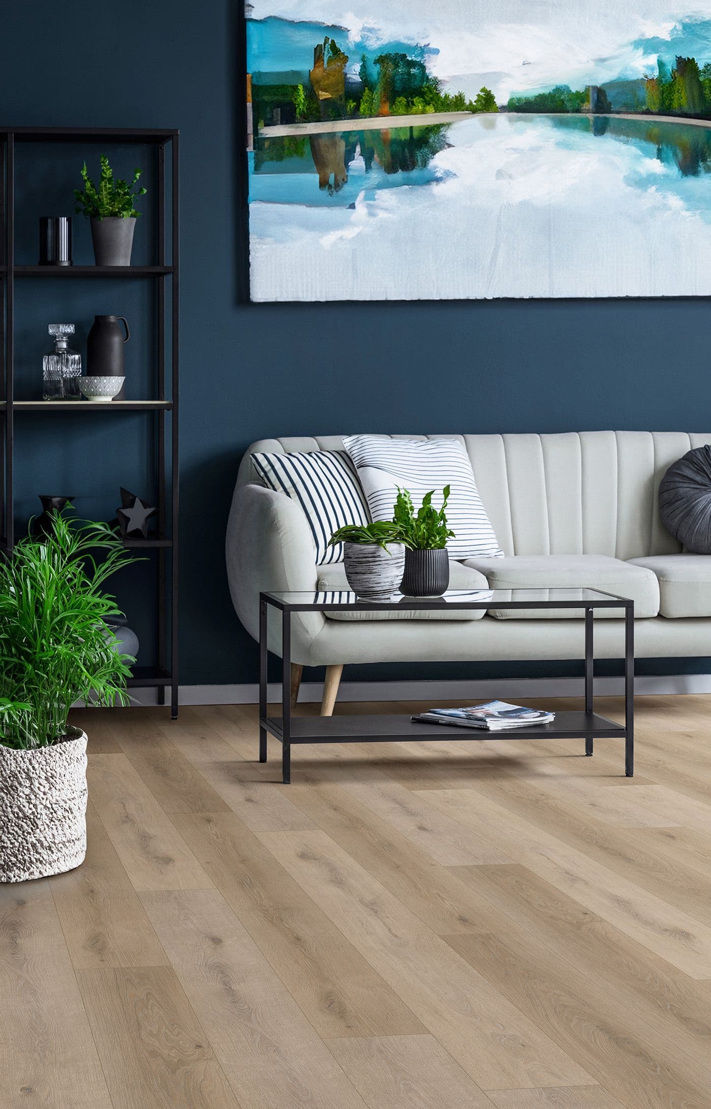Stylish living room with a beige couch, striped pillows, and a black coffee table. A plant-filled shelf and landscape painting enhance the modern vibe.