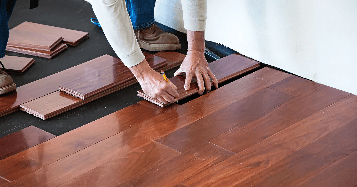 Person installing hardwood flooring, using a pencil to mark cut lines on reddish-brown planks. Tools and stacked planks are visible, conveying focus.