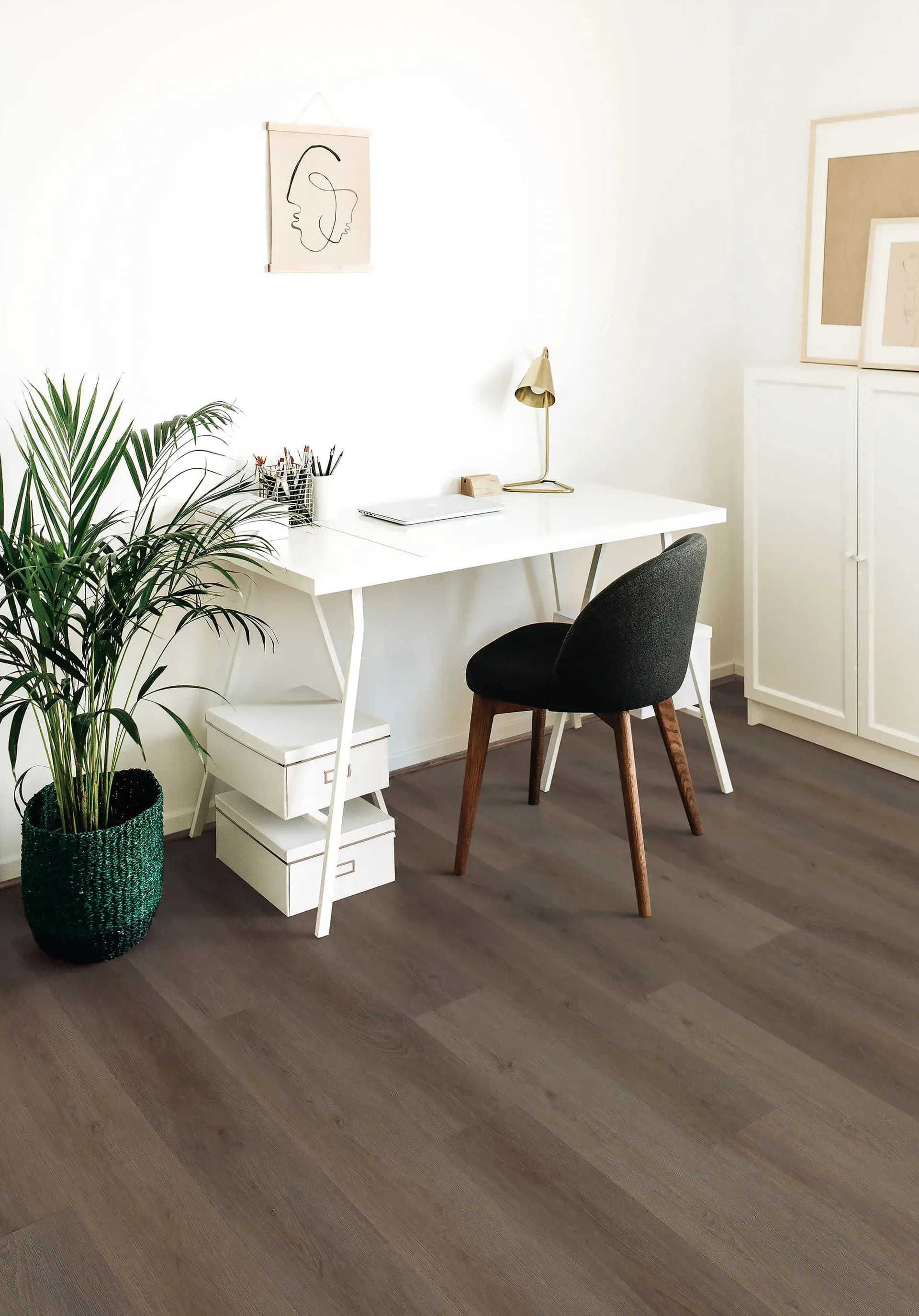 Minimalist home office with a white desk, black chair, gold lamp, and green potted plant. Framed abstracts and storage boxes create an organized vibe.