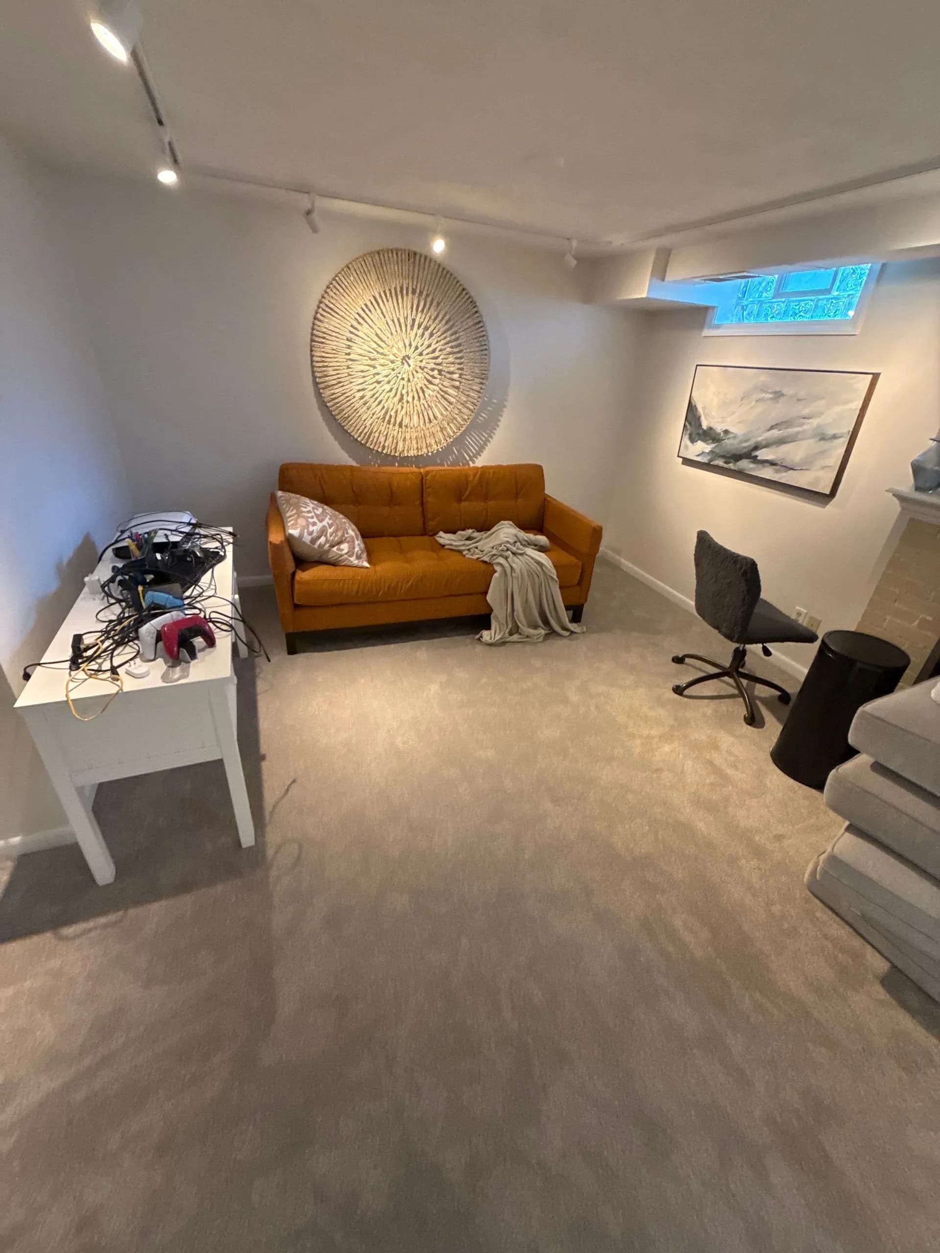 A cozy room with a mustard sofa draped with a blanket, a circular wall art above it, and a small table cluttered with electronics. Soft lighting creates a warm atmosphere.